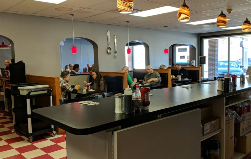 A cozy diner interior with red and white checkered floors, patrons seated at booths, and pendant lights hanging above.