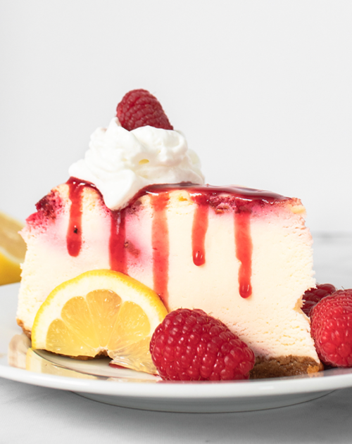 A slice of lemon cheesecake topped with raspberry sauce, whipped cream, and fresh raspberries, served with lemon slices.