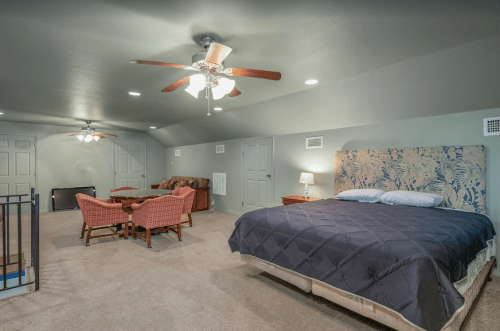 Spacious attic bedroom with a large bed, seating area, and ceiling fans, featuring neutral tones and carpeted floors.
