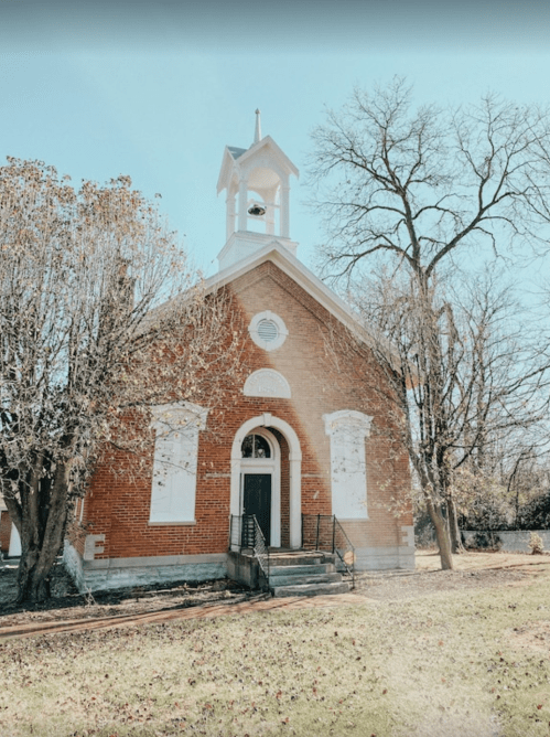 A red brick church with a bell tower, surrounded by trees and a grassy area under a clear blue sky.