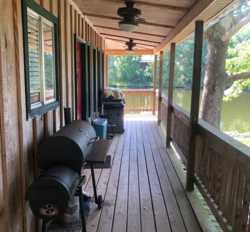 A wooden porch with a grill and a view of a river, surrounded by trees and sunlight.