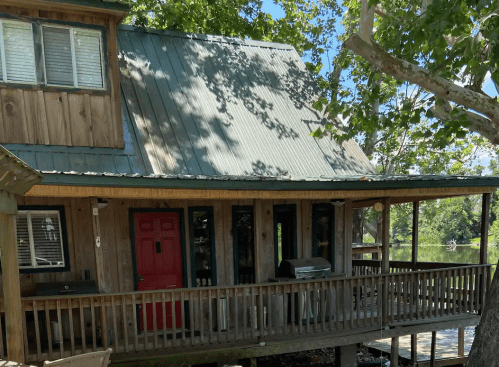 A wooden cabin with a green metal roof, red door, and a porch overlooking a body of water surrounded by trees.