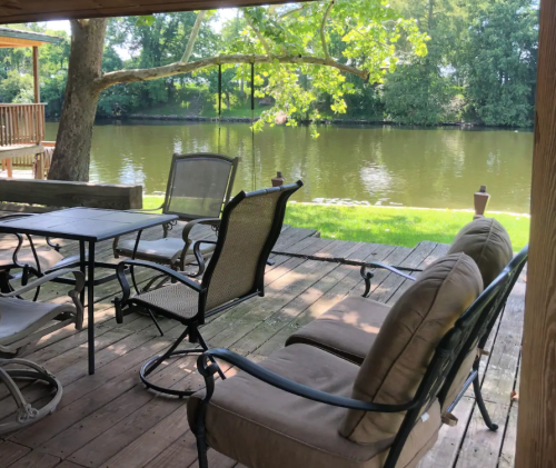 A peaceful riverside patio with chairs and a table, surrounded by greenery and a calm water view.