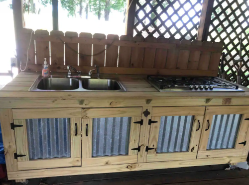 A rustic outdoor kitchen with a wooden countertop, double sink, and a gas stove, featuring metal panel doors.