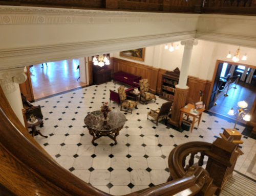 A spacious, elegant lobby with a marble floor, antique furniture, and decorative lighting, viewed from an upper level.