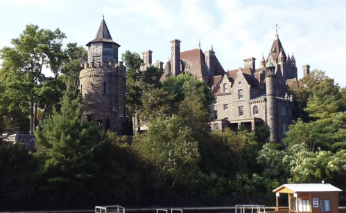 A historic stone castle surrounded by trees, featuring turrets and a tower, set against a clear blue sky.