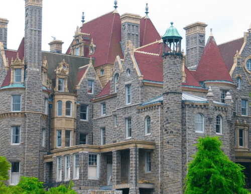 A large, historic stone mansion with red roofs and intricate architectural details surrounded by greenery.