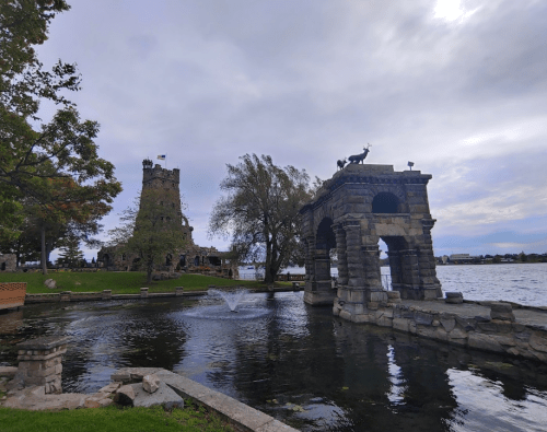 A scenic view of a stone structure and tower by a lake, surrounded by trees and a cloudy sky.