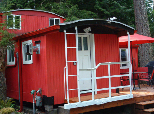 A bright red cabin with a porch, surrounded by trees, featuring a white door and outdoor seating.