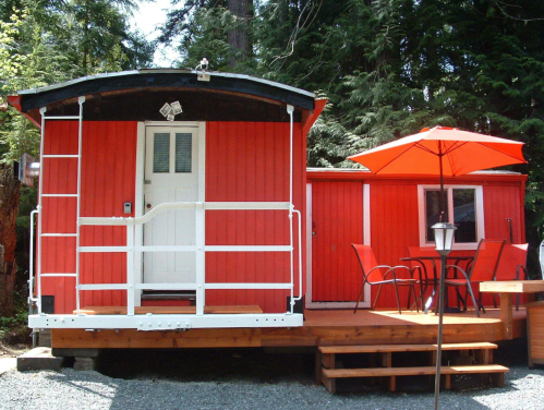 A bright red cabin with a porch, surrounded by trees, featuring a patio with an umbrella and outdoor seating.