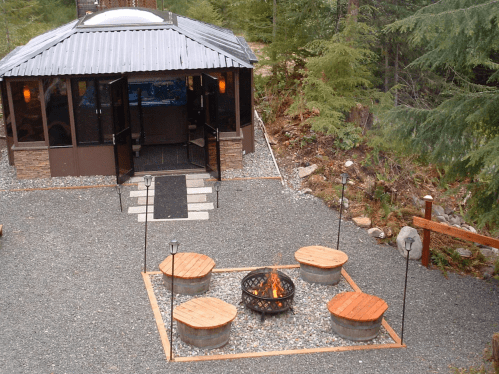 A cozy outdoor space featuring a fire pit surrounded by wooden seating, with a cabin in a forested area.