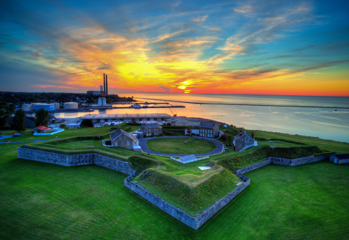 A scenic view of a historic fort at sunset, overlooking a calm bay with buildings and a smokestack in the background.