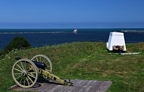 A historic cannon on a grassy hill overlooking the ocean, with a lighthouse visible in the distance under a clear blue sky.