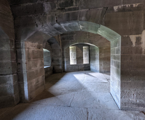 Interior of a stone structure with arched openings and natural light illuminating the floor.