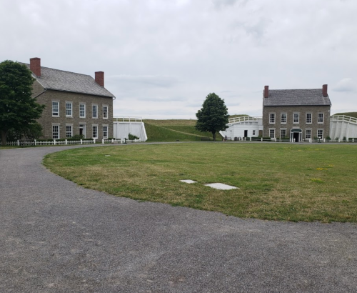 Two historic buildings with gray stone exteriors, surrounded by a grassy area and a gravel path under a cloudy sky.