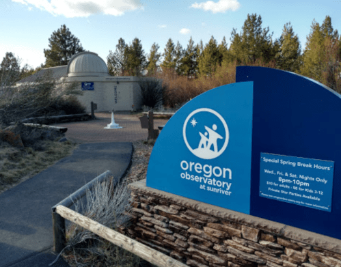Sign for the Oregon Observatory at Sunriver, with a dome building in the background and a pathway leading up to it.