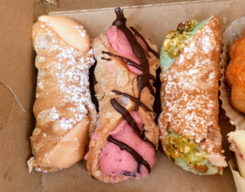 Three colorful cannoli filled with cream, dusted with powdered sugar, in a cardboard box.