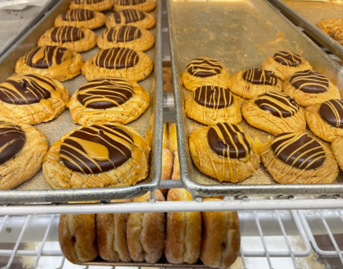 Trays of cookies topped with chocolate and drizzled with caramel, alongside a row of donuts.