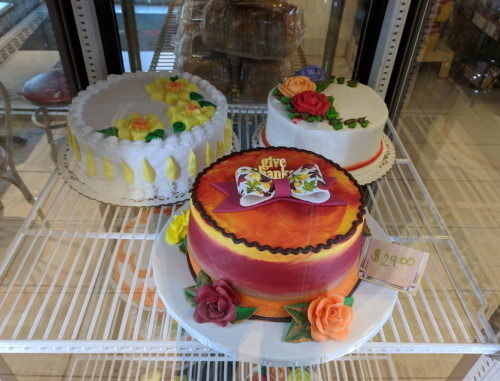 Three decorative cakes in a display case, featuring flowers and a "give thanks" bow on the center cake.