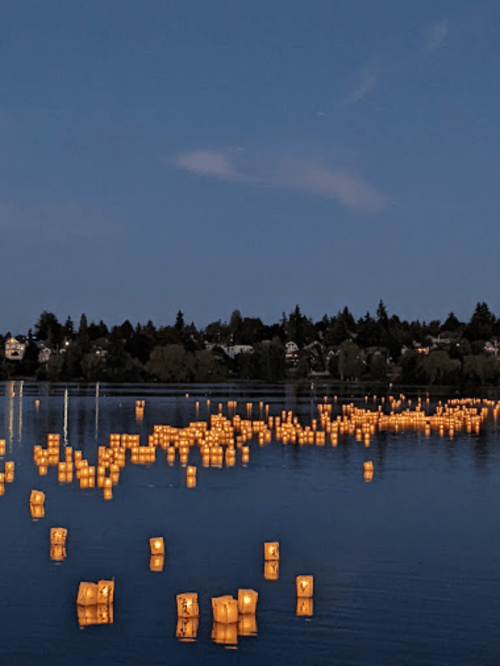 Floating lanterns illuminate a calm lake at dusk, surrounded by trees and houses under a twilight sky.