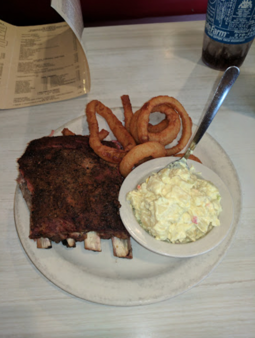 A plate with ribs, onion rings, and a side of potato salad on a white table.