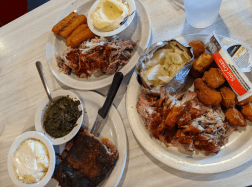 Two plates of barbecue with pulled pork, ribs, sides of greens, mashed potatoes, and cornbread, served with a drink.
