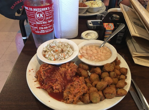 A plate of pulled pork with sauce, fried okra, a side of rice, and white beans, served with a drink and napkin.