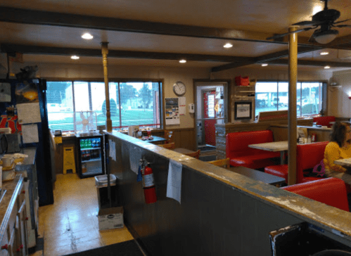 Interior of a diner with red booths, a counter, and large windows showing an outdoor view. A clock and fan are visible.