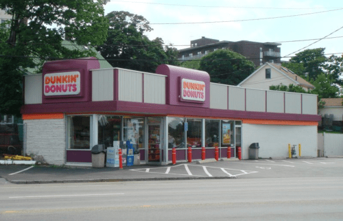 A Dunkin' Donuts store with a purple and white exterior, featuring large signage and a parking area in front.