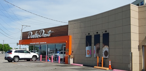 Exterior of a donut shop with a sign, large windows, and a parking area, featuring colorful donut images on the wall.