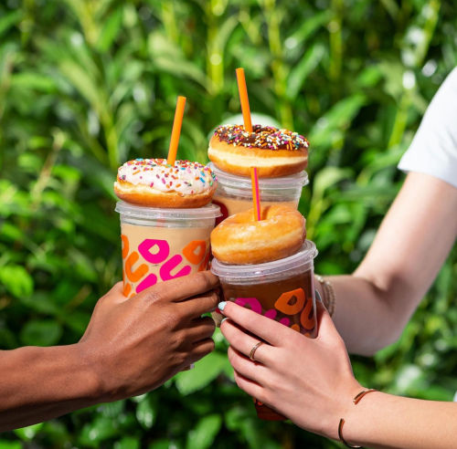 Four hands holding colorful drinks topped with donuts, set against a lush green background.
