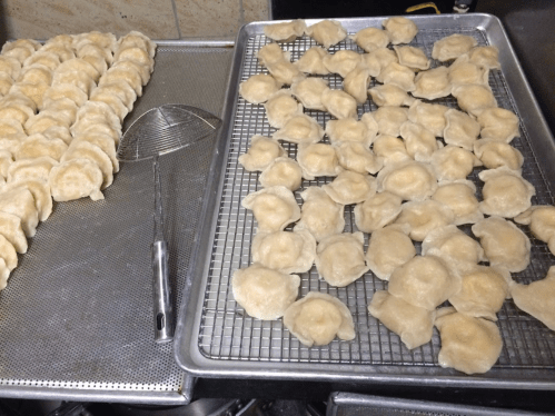 A tray of uncooked dumplings arranged neatly on a metal rack, with a slotted spoon nearby.