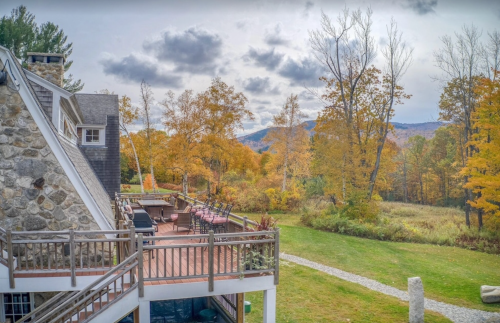 A scenic view of a house with a deck, surrounded by autumn trees and mountains under a cloudy sky.
