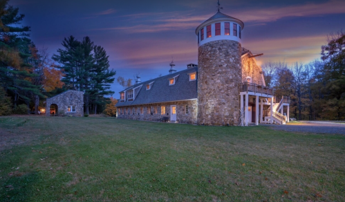 A stone building with a circular tower, surrounded by trees and a grassy area, under a colorful sunset sky.