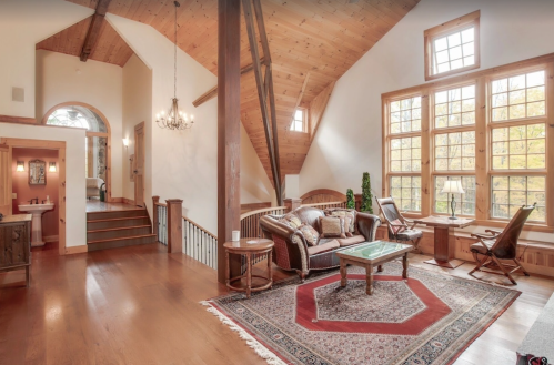 Cozy living room with wooden beams, large windows, and a patterned rug, featuring comfortable seating and warm lighting.
