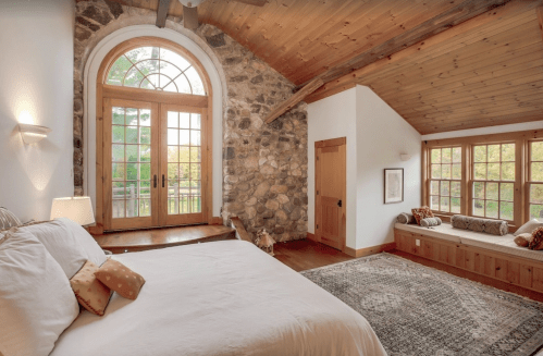 Cozy bedroom with stone walls, large windows, and wooden accents, featuring a bed and a window seat.