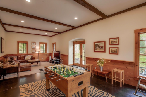 A cozy game room featuring a foosball table, a sofa, and large windows with natural light.