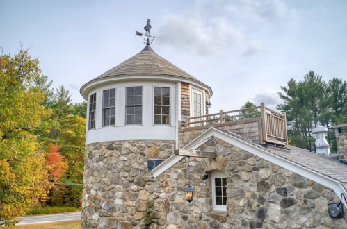 A stone building with a circular tower, featuring a weather vane, surrounded by trees in autumn colors.