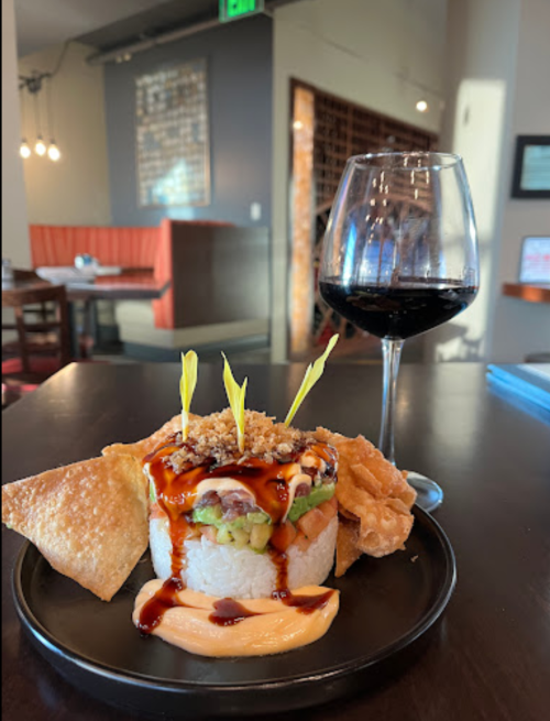 A plate of sushi topped with sauce and garnishes, accompanied by crispy chips and a glass of red wine.