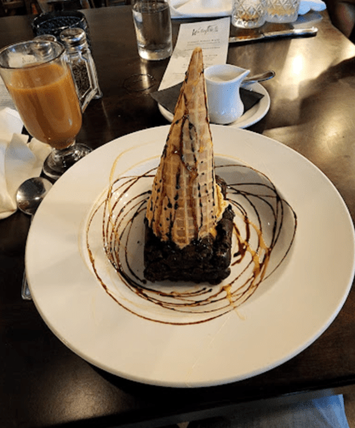 A dessert featuring a brownie topped with a cone of ice cream, drizzled with chocolate sauce on a white plate.