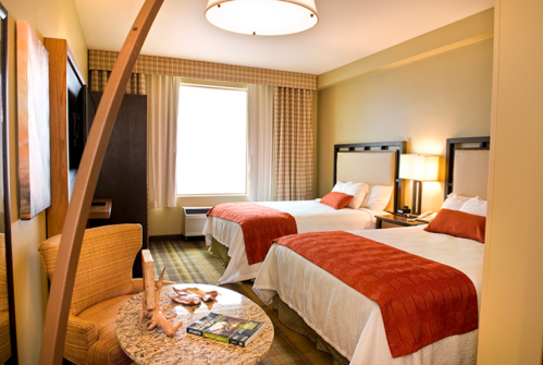 A cozy hotel room featuring two beds with orange accents, a small table, and a large window with natural light.