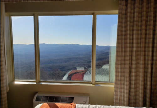 A cozy room with a bed and large windows showcasing a scenic mountain view under a clear blue sky.