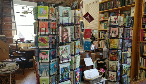 A cozy bookstore interior with colorful greeting cards displayed on racks and bookshelves in the background.