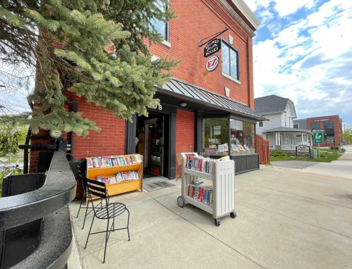 A cozy bookstore with outdoor book displays, surrounded by greenery and residential buildings under a partly cloudy sky.