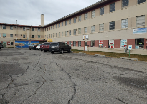 A parking lot in front of a large, beige building with murals on the walls and cloudy skies above.