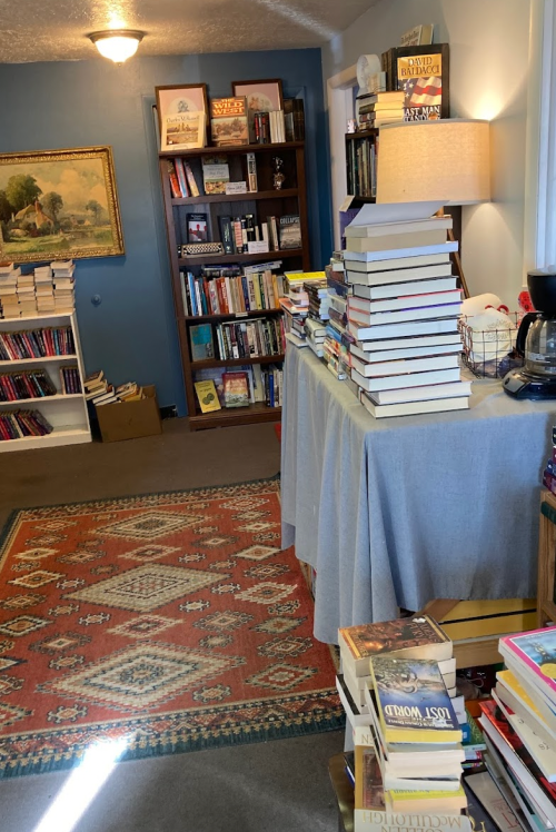 A cozy room filled with bookshelves, stacks of books, and a warm rug, creating a welcoming reading space.