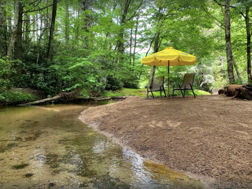 A serene spot by a creek with two chairs under a yellow umbrella, surrounded by lush green trees.