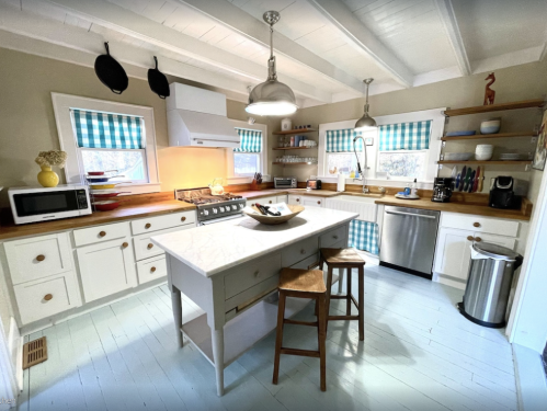 Bright kitchen with white cabinets, a central island, wooden shelves, and blue checkered curtains. Modern appliances included.
