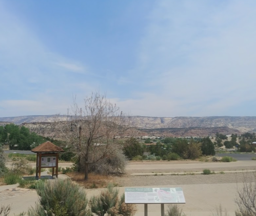 A scenic view of a desert landscape with a sign, sparse vegetation, and distant cliffs under a blue sky.