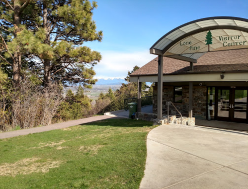 A visitor center with a curved roof, surrounded by trees and a view of distant mountains under a clear sky.